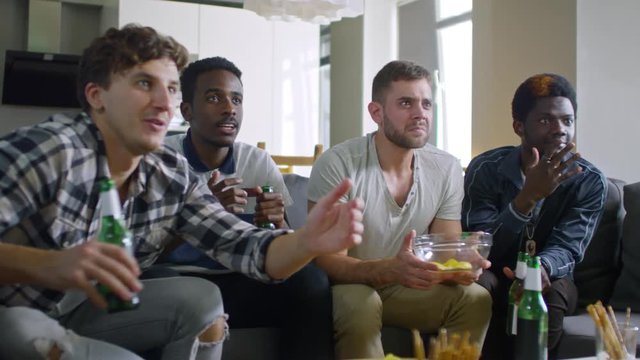 Multi-ethnic group of young men sitting together on couch, drinking beer and getting very upset about failure of soccer team