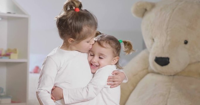 Portrait of two sisters in their bedroom smiling and kissing, in the background the stuffed animals and the dollhouse. Concept of: family, sisters, home and happiness.