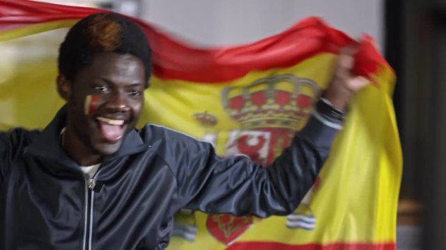 Young African-American man smiling and dancing with flag of Spain while sitting on sofa and watching TV match at home