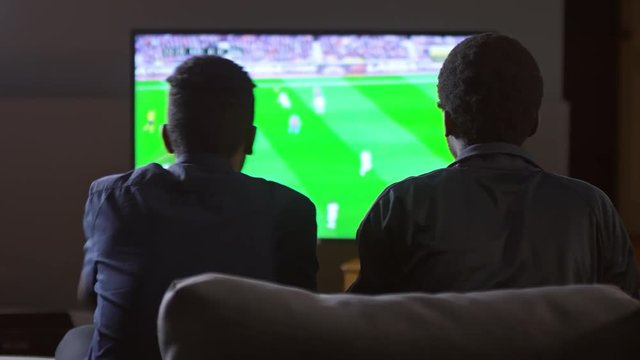 Rear view of two African-American men sitting on couch at home, talking and discussing game while watching soccer on TV