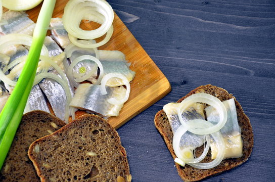 Salted herring slices with onion rings and greens in oil with spices and a sandwich on the table and with pieces of rye bread with seeds