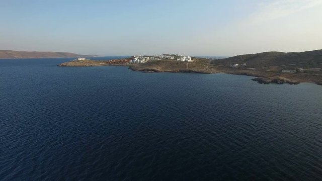 Flying over waters of Aegean Sea towards peninsula with stony shore, hills covered with dry grass, cobblestones, dusty roads and small settlement with white villas on top. Attica, Greece. Aerial.