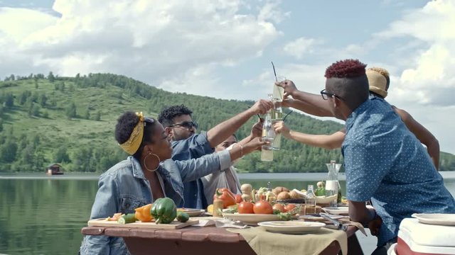 Group of six young people sitting at table during picnic by lake on summer day and clinking bottles