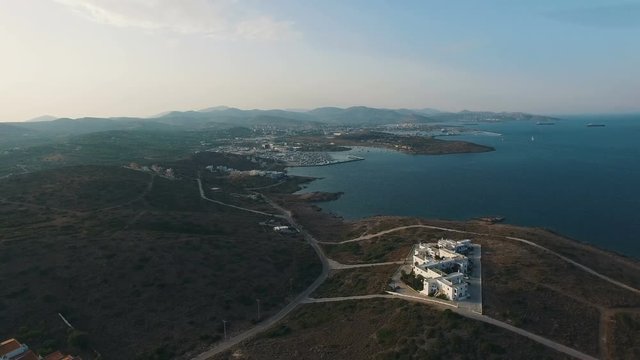 Flight over road along shore of Aegean Sea at sunset. Desolated white villas and buildings on hills covered by arid bushes, shrubs. Big port city and mountains on background. Attica, Greece. Aerial