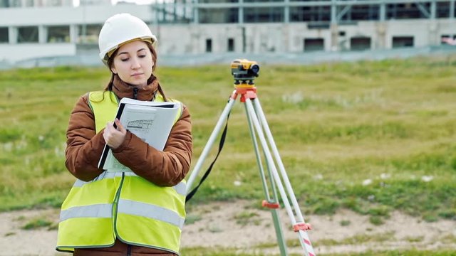 A young woman surveyor in work clothes and helmet adjusts the equipment and produces calculations on the construction site. The concept of landscape design, geodesy