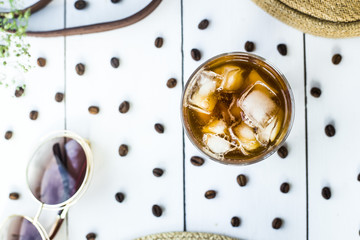 Cold coffee in a glass with ice. On the table among coffee beans and summer women's accessories. Summer relax concept. Flat lay, top view