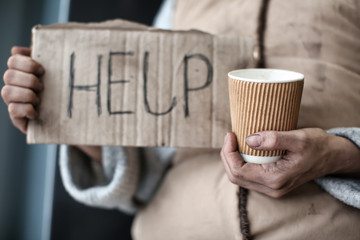 Poor woman holding piece of cardboard with word HELP and cup, closeup