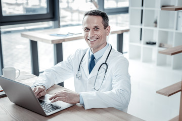 Ready to work. Satisfied confident handsome doctor sitting on his workplace smiling and using the laptop.