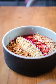 Dragon fruit smoothie bowl with banana, strawberries, granola, coconut milk from above. Wooden table at coffee shop. Minimalist food photography