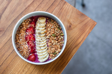 Overhead dragon fruit smoothie bowl with banana, strawberries, granola, coconut milk. Wooden table at coffee shop. Minimalist food photography