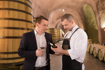 Portrait two men tasting with red wine in a vineyard. Professional winemakers standing in front of wine cellar and consulting.
