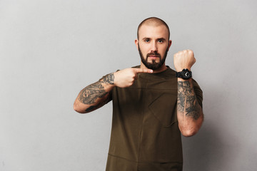 Portrait of serious baldy guy in casual t-shirt looking on camera and pointing finger on his wrist watch, isolated over gray background