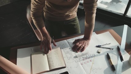 Hipster engineer works with blueprint. Shoot from above. Young architectural engineer working in office. Bearded man in glasses makes sketches. Loft style, minimalistic interior, drawings on the table
