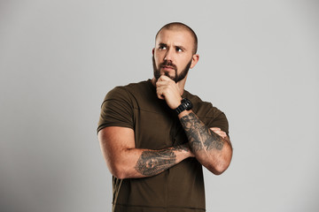 Portrait of manly bald guy with tattoo on his arms looking aside and holding his chin, isolated over gray background