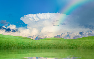 Colorful rainbow above green hills, blue sky with white incus cloud, serene lake