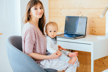 Charming european woman working with laptop and holding infant baby boy at home interior....