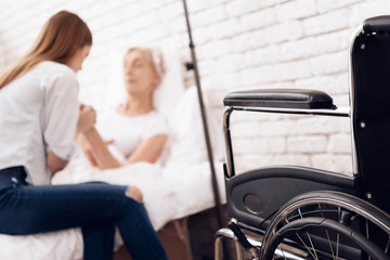 Girl is nursing elderly woman at home. They are holding hands. Wheelchair is standing near.