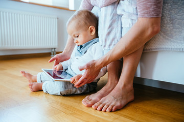 Cute infant baby boy barefoot using a tablet sitting on the flloor, his unrecognizable mother helping him to hold gadget. Real people life concept.