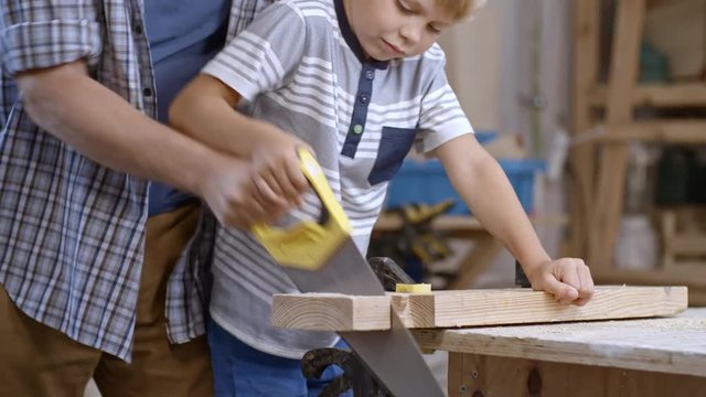 Boy of primary school age learning to use hand saw to cut wooden block, his father helping him