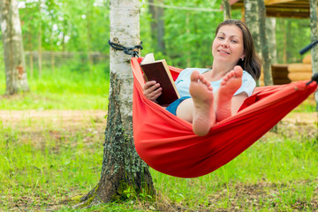 Naklejka premium happy 30 year old woman reading a book, sitting in a red hammock in the garden
