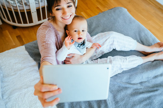 Mother and baby with funny faces photographing selfie themselves by mobile tablet at home interior.