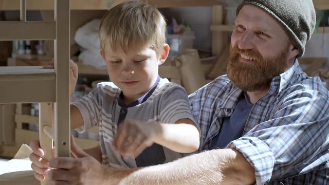 Boy of primary school age working in art studio and making chair of wood with help of his father