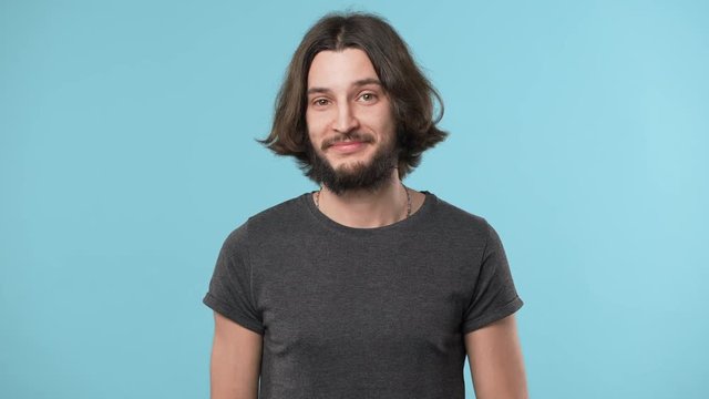 Portrait of hairy man wearing casual gray t-shirt grinning sarcastically and expressing distrust or disbelieve, over blue background. Concept of emotions