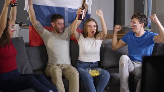 Russian soccer fans with flag paint on their cheeks sitting on couch and watching TV game. Young man holding flag of Russia and women toasting with beer