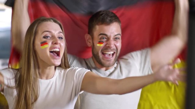 Beautiful female soccer fan and her boyfriend holding flags of Spain, watching TV match, kissing, shouting and celebrating goal