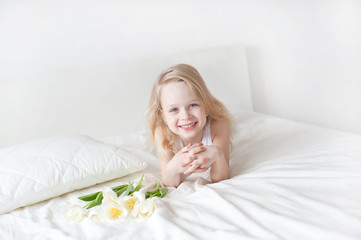 Obraz na płótnie Canvas Beautiful little girl looking at camera and smiling. Girl child on a big white bed in a white room.