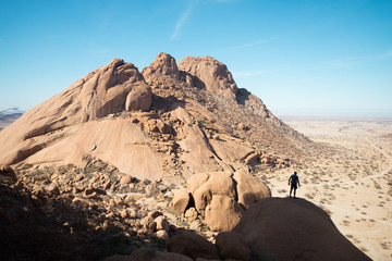 A single hiker silhouetted against the harsh desert and rocky peaks of Namibia having climbed up a mountain