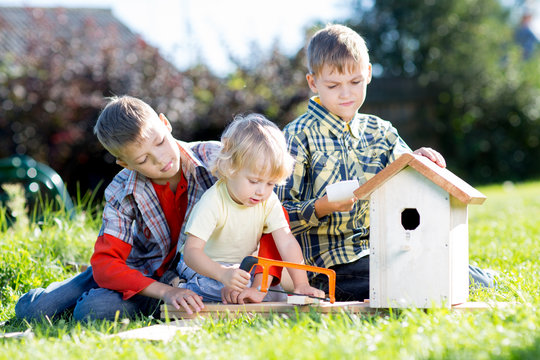 Kids Boys Making Birdhouse Together Sitting On The Grass. Oldest Child Teaches Youngest Brother To Work With Tools