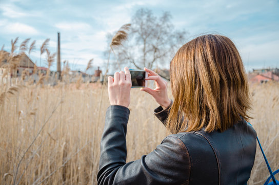 Young brunette woman taking photo in bulrush in nature with her phone, wearing leather jacket