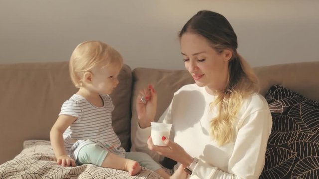 Young mom feeding little daughter with yogurt on sofa. Little girl eating yogurt