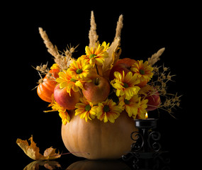 Bouquet of apples, flowers and dried plants in vase from pumpkin next to candle is burning, isolated on black