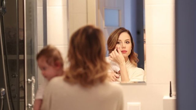 mother holding a litle baby and appying cosmetics