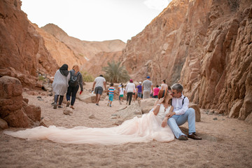 Bride and groom sit and kiss in canyon against the backdrop of tourists passing by.