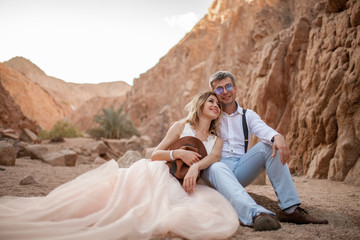 Bride and groom sit and smile in canyon on sand on background of rocks.