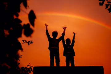 Two brothers with their hands up under the rainbow on sunset in summer.