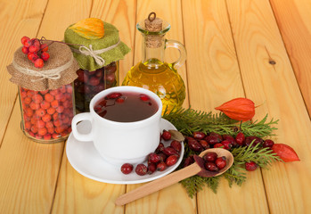 Banks with raw materials, jug of a rosehips oil, a drink in cup and scattered rosehip, on wooden table