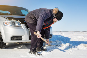 The driver digs out the car with a shovel from the snow. The car is stuck in the snow, the man shovels the snow from under it.