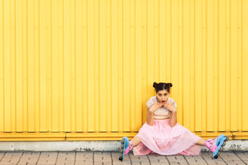 young girl in pink skirt and roller skates sits on the sidewalk against the background of yellow wall. Emotions with an open mouth. Concept life style. Copy space