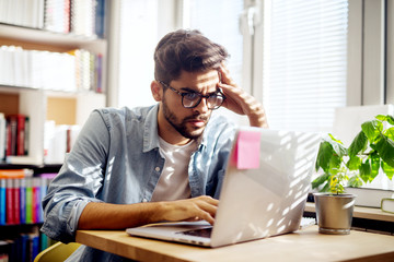 Handsome bearded young student guy looking confused at a laptop while sitting at the library.