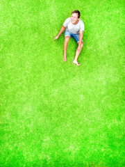 A man lies on a green lawn and smiles with happiness. Top and aerial view with copy space. Minimal styled flat lay isolated on original green grass background. Sits and look up.