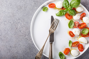 White plate of healthy classic delicious caprese salad with ripe tomatoes and mozzarella cheese with fresh basil leaves on gray concrete background with space for text. Italian food. Top view.