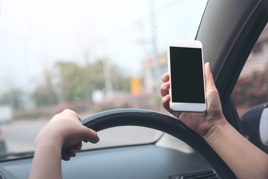 Mockup image of a hand holding and using white mobile phone with blank black desktop screen while driving car on the road