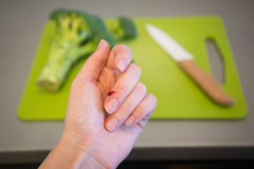 Wounded finger with blood and knife on cutting board with broccoli on grey background