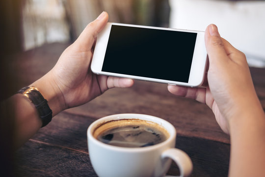 Mockup image of hands holding white mobile phone with blank black screen for watching and playing games with a cup of coffee on wooden table in cafe