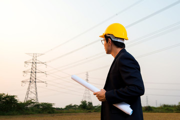 An engineer wear yellow hard hat and suite hold blueprint in hand standing on field looking at electricity towers in background