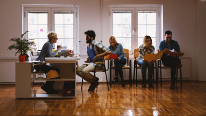 Side view of group young people sitting and waiting in the line with a portfolio for a job interview while one of them having a conversation with the entrepreneur.